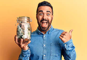 Delighted man holding a huge jar of cash in one hand while giving the thumbs up with the other