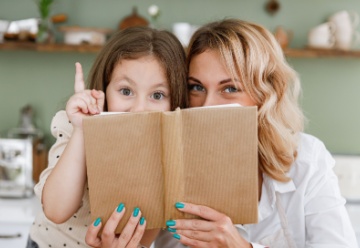 Mom woman in white shirt work baby girl helper read cookbook point finger up hide at kitchen table home