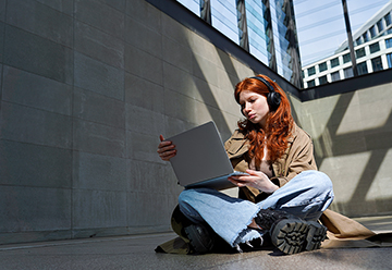 A young woman wearing headphones is sitting cross-legged on the floor looking at a laptop and wearing headphones