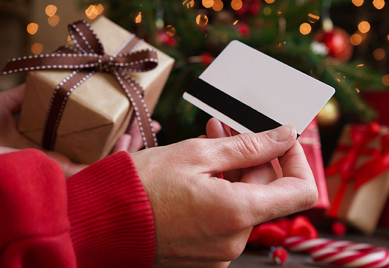 Young woman holding a credit card and a gift box against the background of Christmas decor and gifts close-up. Christmas and New Year shopping on the Internet. Payment by gift card