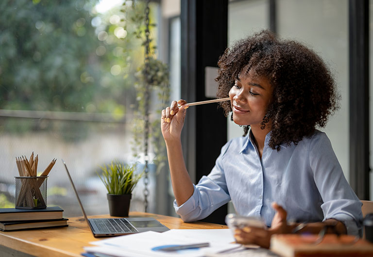 Smiling woman sitting at a desk with a laptop, paperwork on it and a pen in her hand